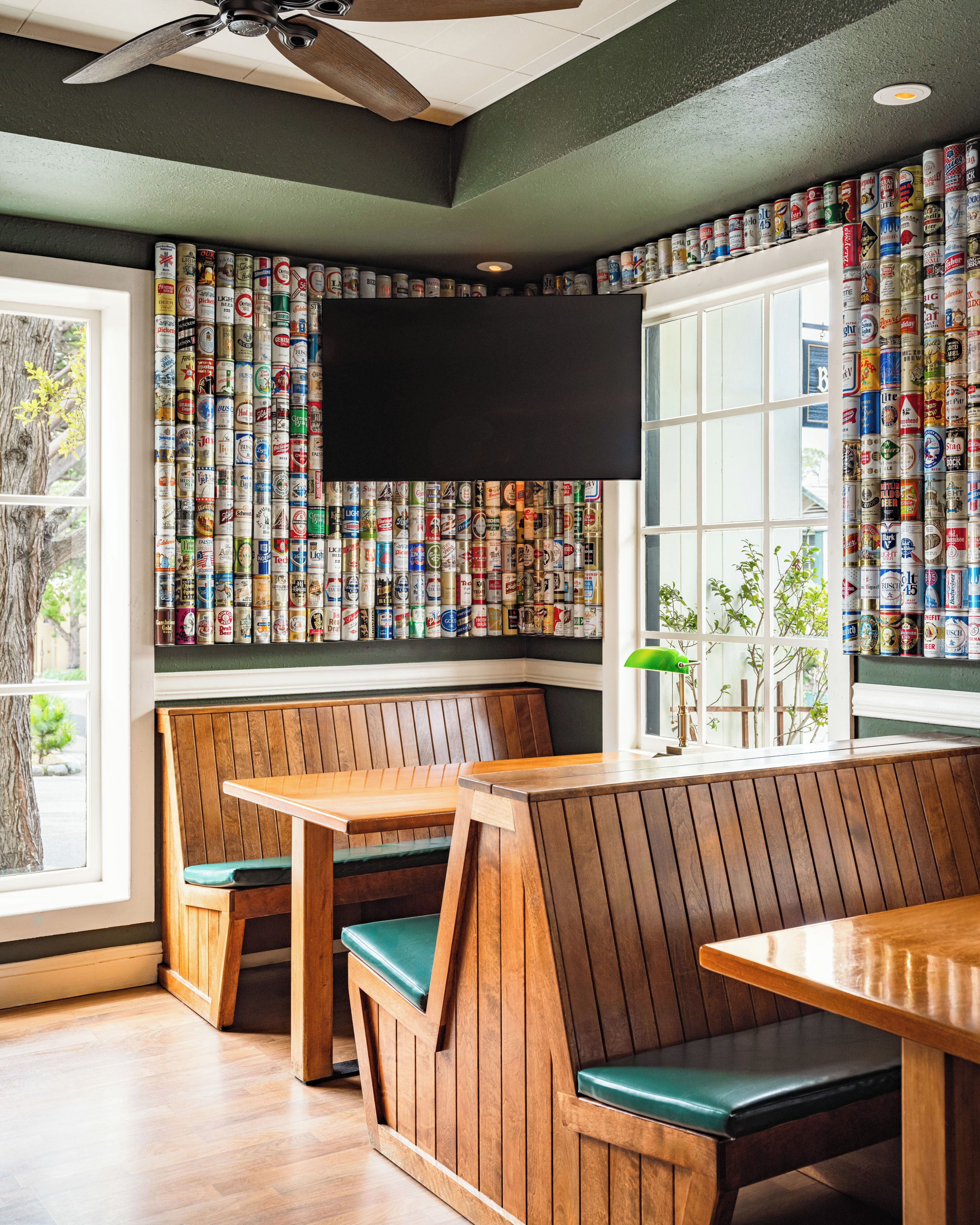 A restaurant interior with wooden benches and tables, a wall of stacked beer cans, and a flat-screen TV mounted above the seating area.