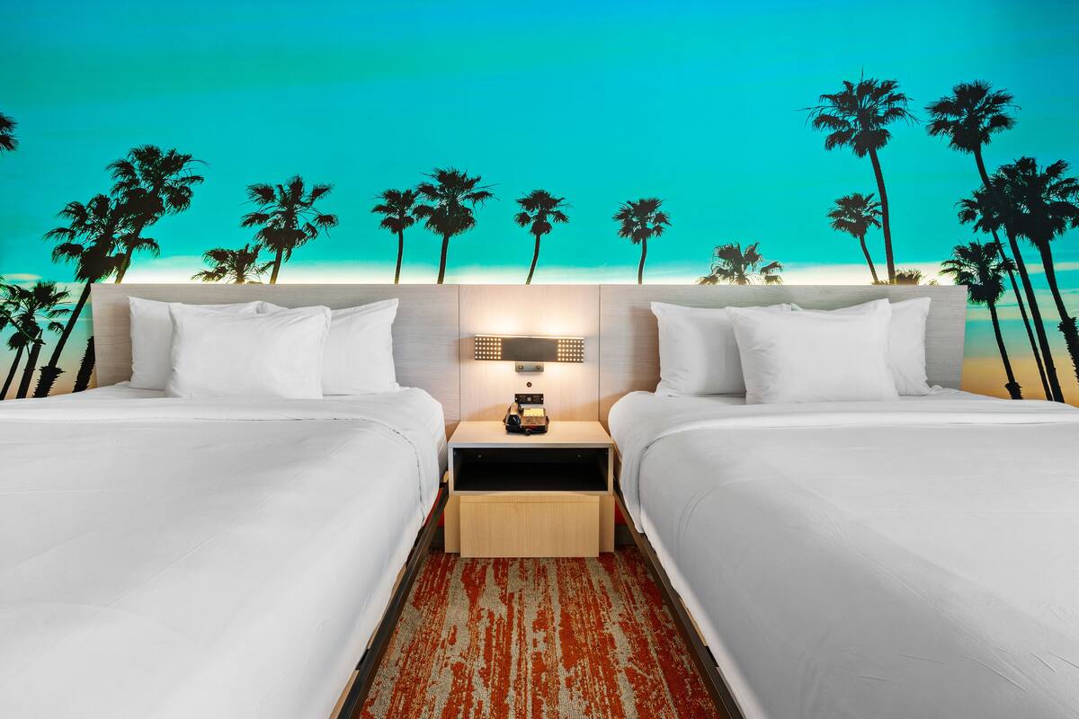 A hotel room with two beds, a nightstand with a lamp, and a view of palm trees.