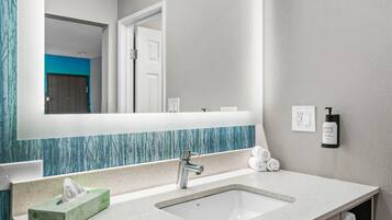 Un baño con un lavamanos blanco, un espejo, un perchero para toallas y una botella de gel antibacterial.