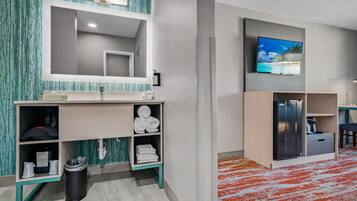Une salle de bain moderne avec un grand miroir, un lavabo, des toilettes et un espace douche. Un téléviseur est fixé au mur, et une petite étagère contient des serviettes.