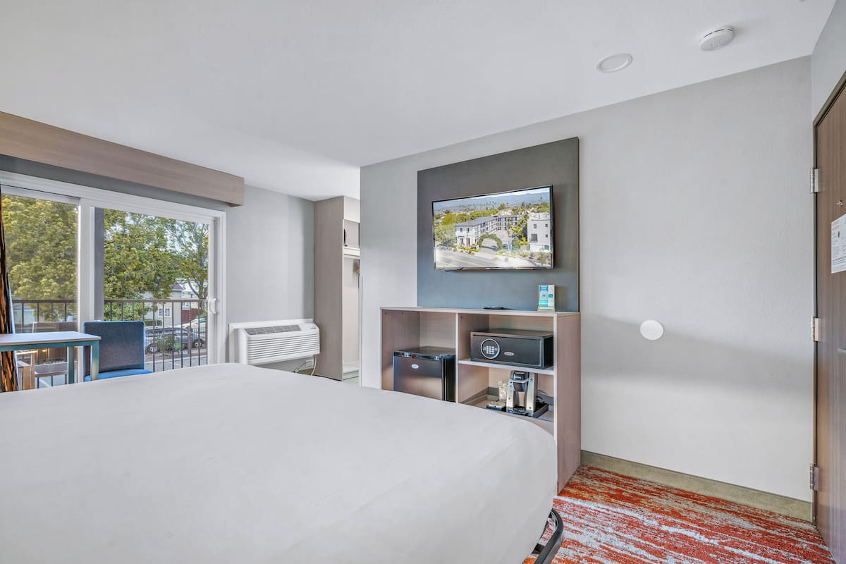 A hotel room with a large bed, a television mounted on the wall, a small desk with a chair, and a view of the outside through a window.