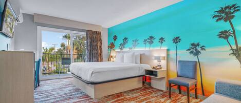 Quarto de hotel moderno com uma cama grande, uma escrivaninha, uma cadeira e vista para palmeiras através de uma varanda.