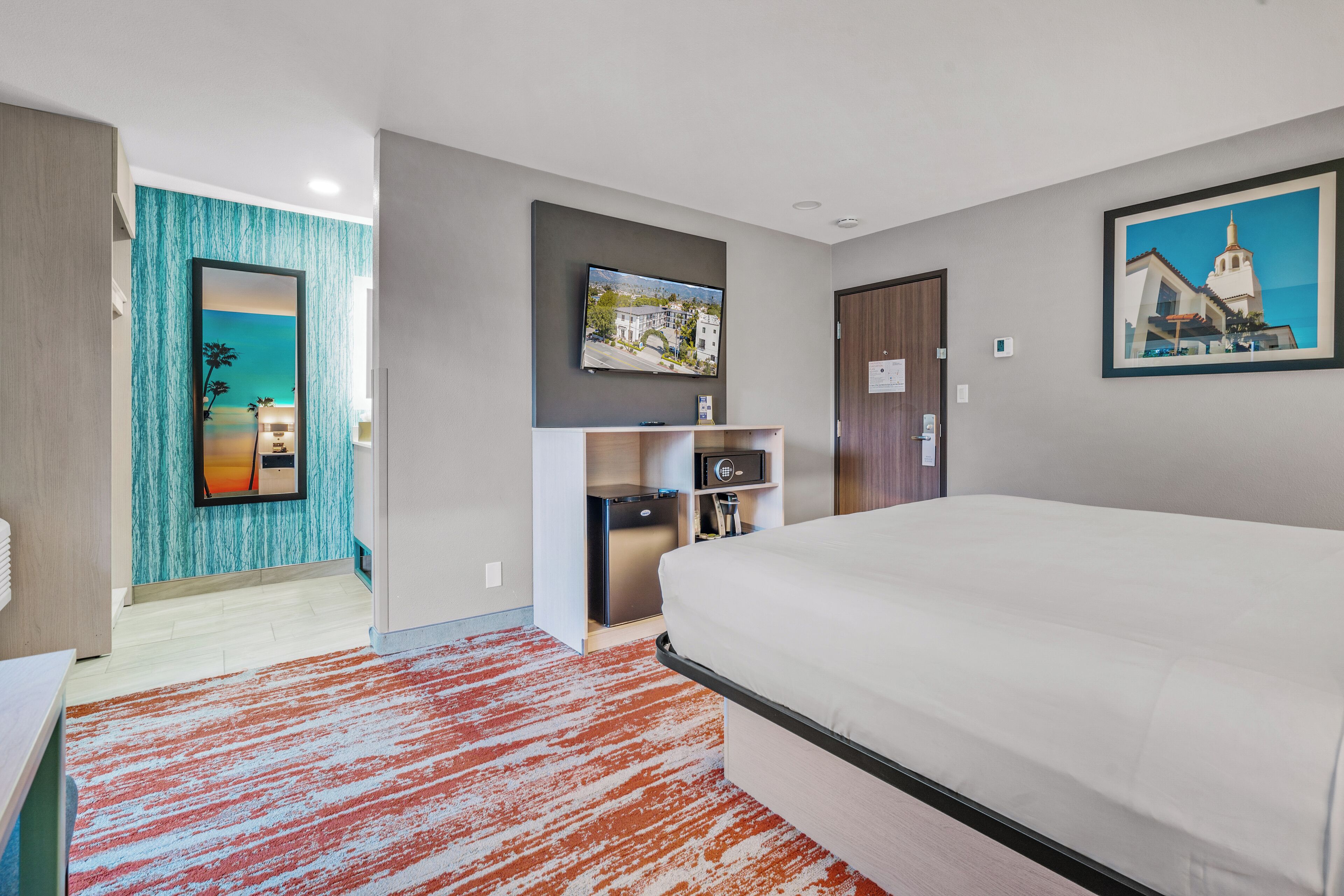 A modern hotel room with a large bed, a flat-screen TV mounted on the wall, a small desk with a chair, and a view of a building through the window.