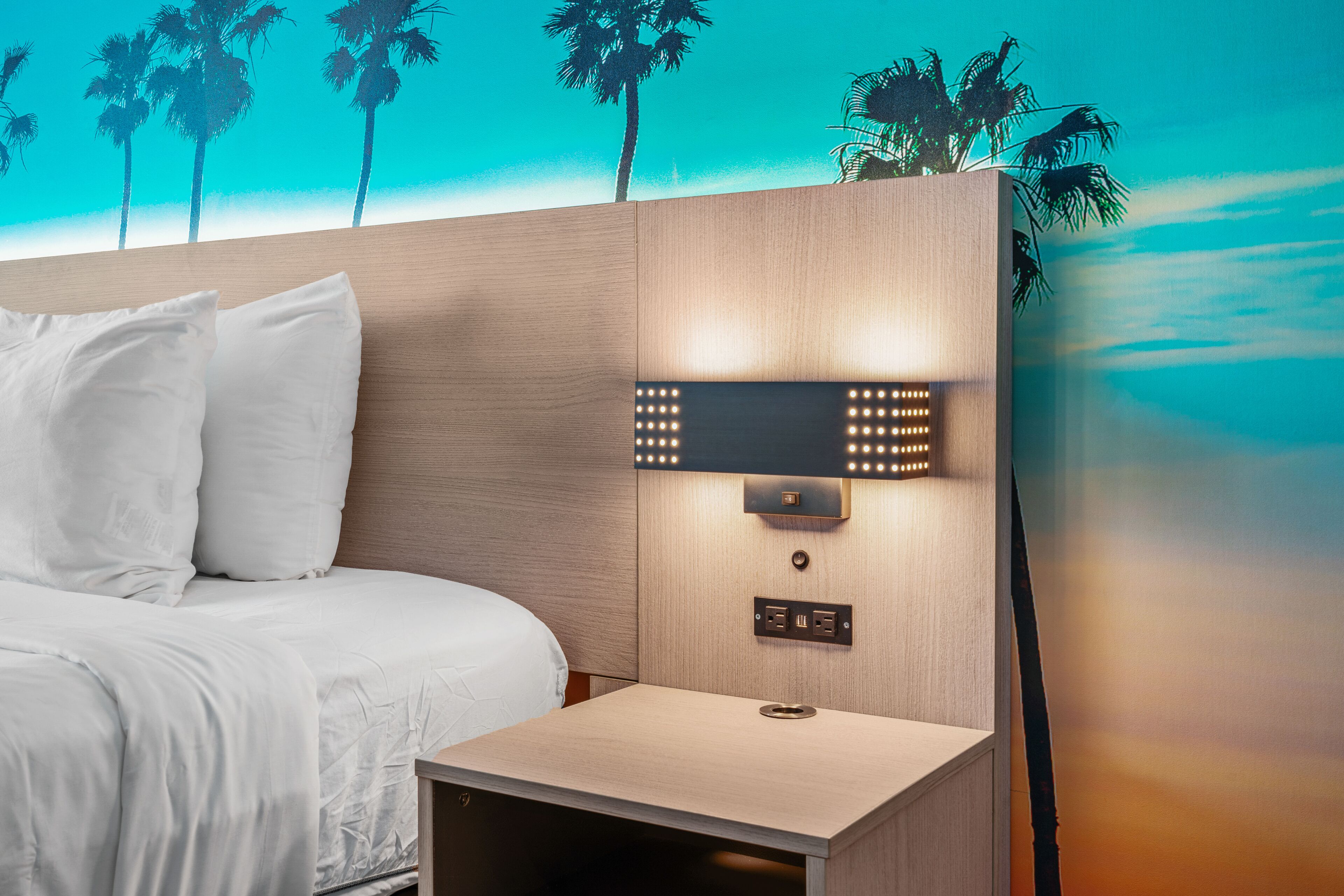 A hotel room with a bed, a nightstand, a wall-mounted light fixture, and a wall mural of palm trees.