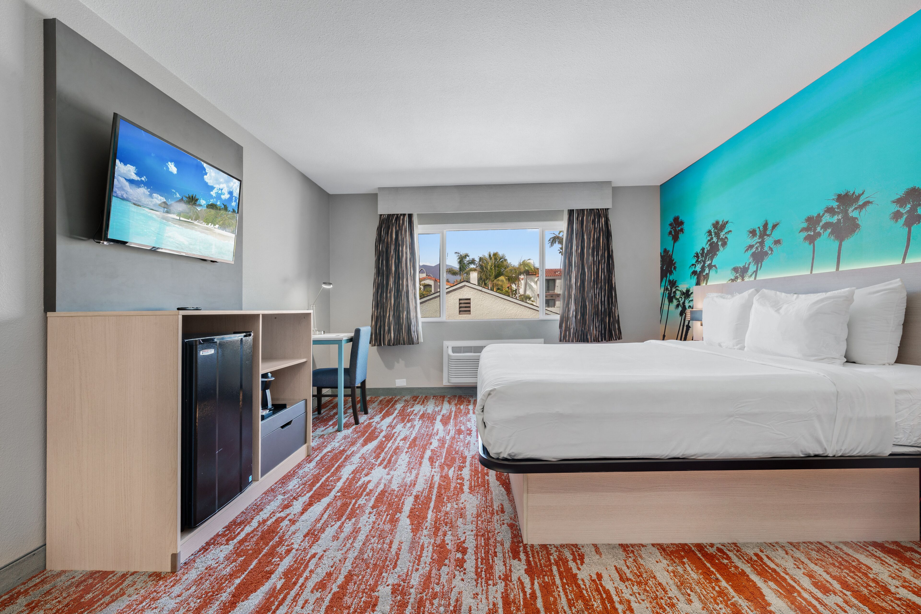 A hotel room with a bed, a desk, a TV, and a view of palm trees and a beach.