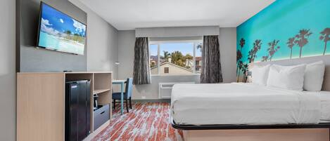 A hotel room with a bed, a desk, a TV, and a view of palm trees and a beach.