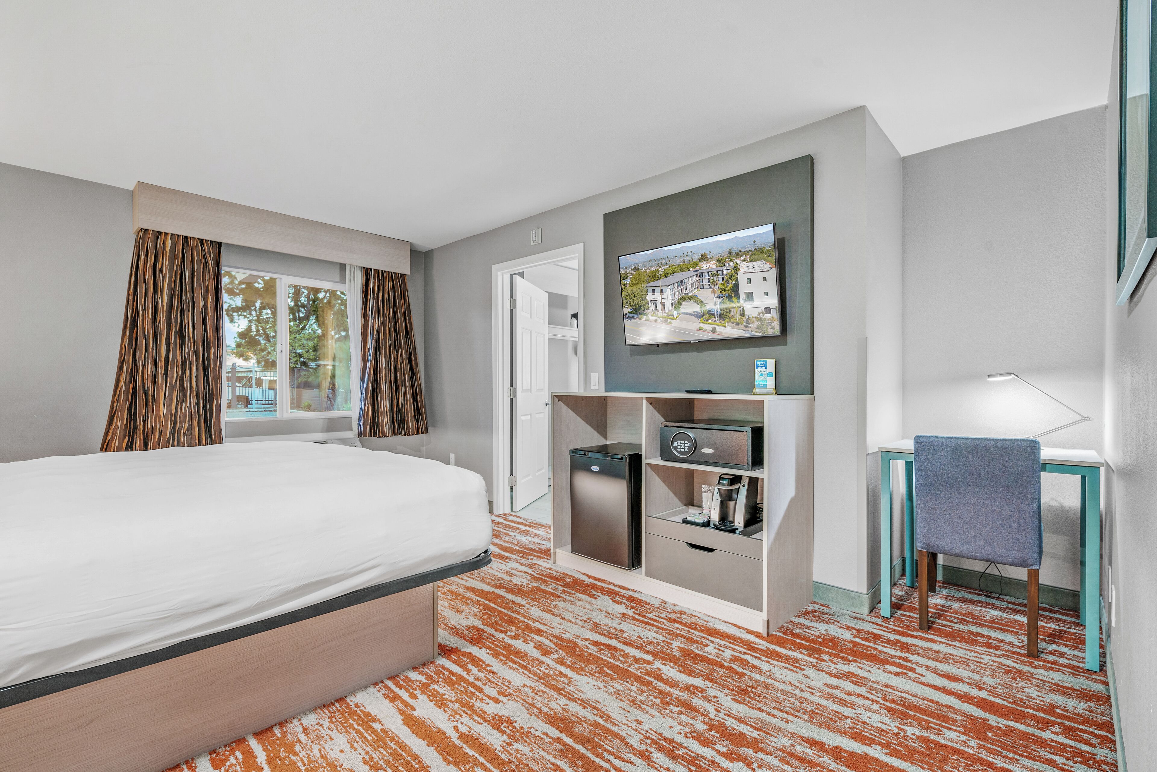 Quarto de hotel com uma cama, uma secretária com cadeira, televisão na parede e uma pequena área de cozinha.