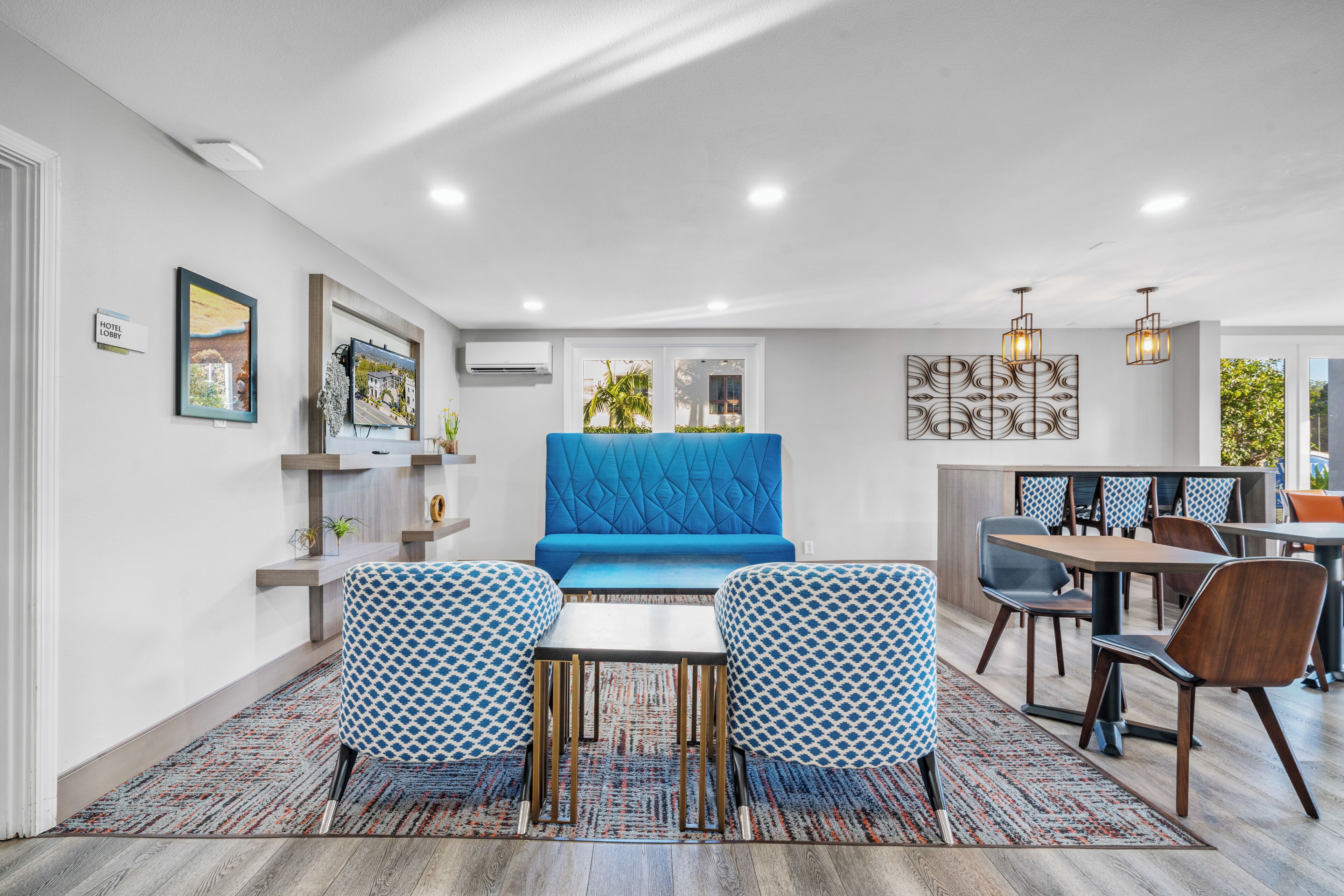 Una habitación de hotel moderna con un sofá azul, sillones estampados, una zona de comedor con sillas de madera y un adorno decorativo en la pared.