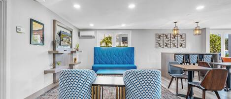 Una habitación de hotel moderna con un sofá azul, sillones estampados, una zona de comedor con sillas de madera y un adorno decorativo en la pared.