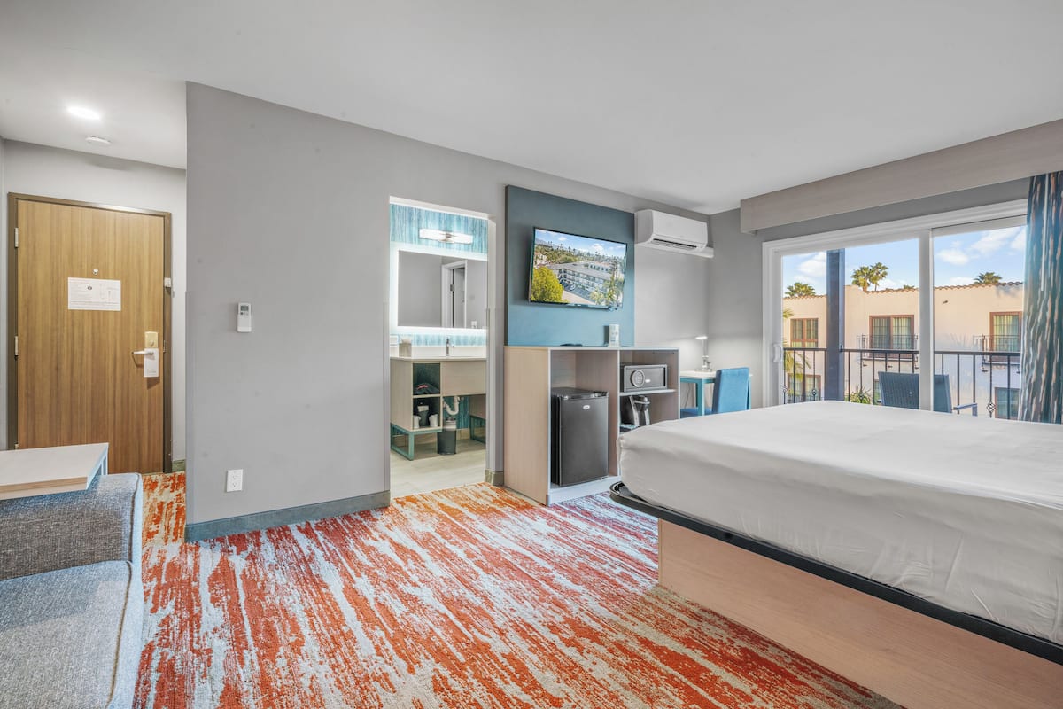 A hotel room with a bed, a desk, a chair, a TV, a mini-fridge, and a bathroom with a shower and toilet.