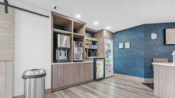 A modern kitchen with wooden cabinets, a stainless steel trash can, a microwave, and a refrigerator with a blue patterned wall behind it.