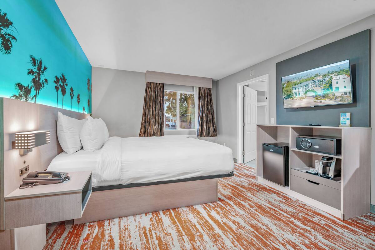 A modern hotel room with a large bed, a TV, a mini-fridge, and a desk with a lamp.