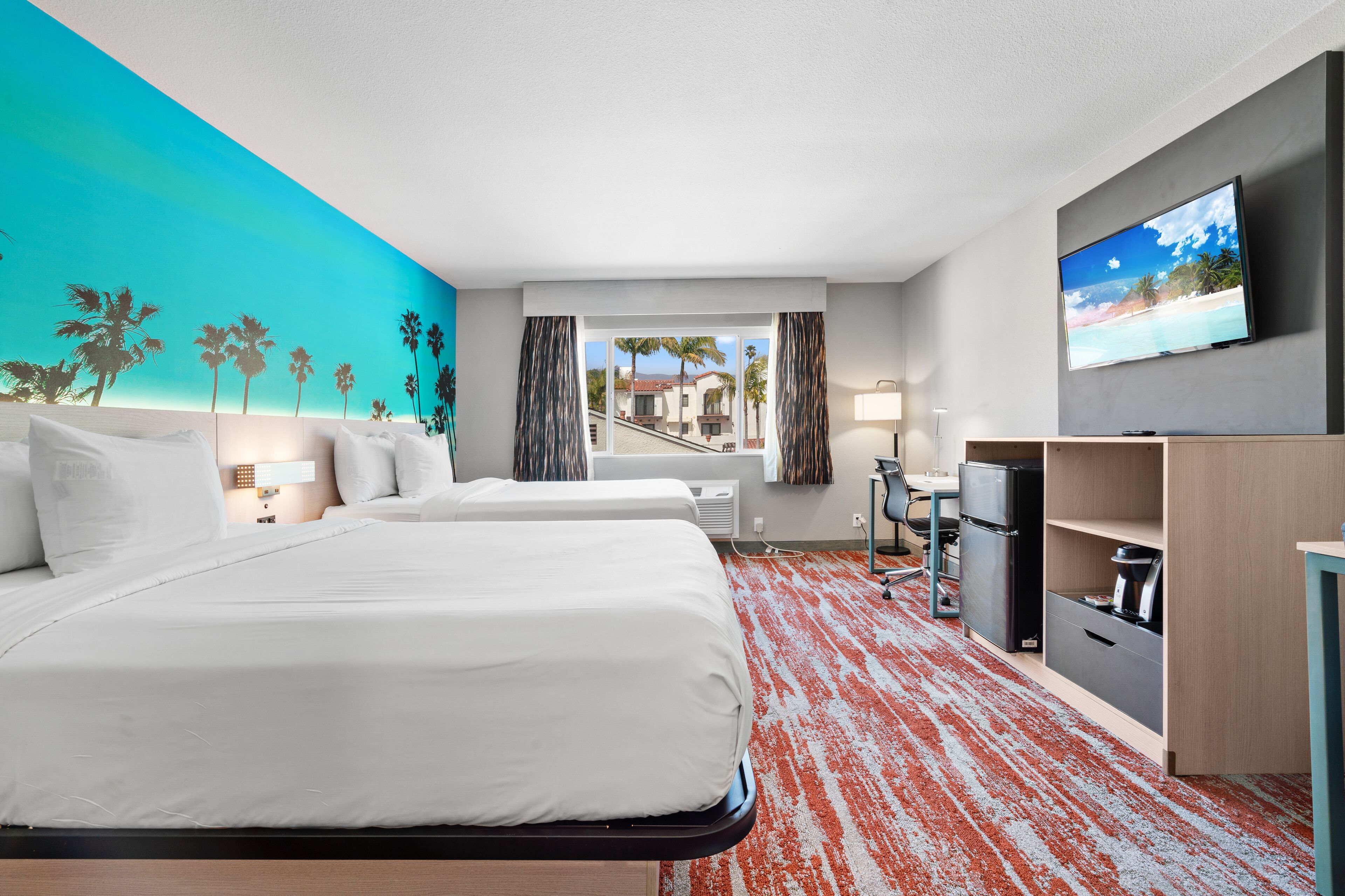 A hotel room with two beds, a TV, a desk, and a view of a beach through the window.