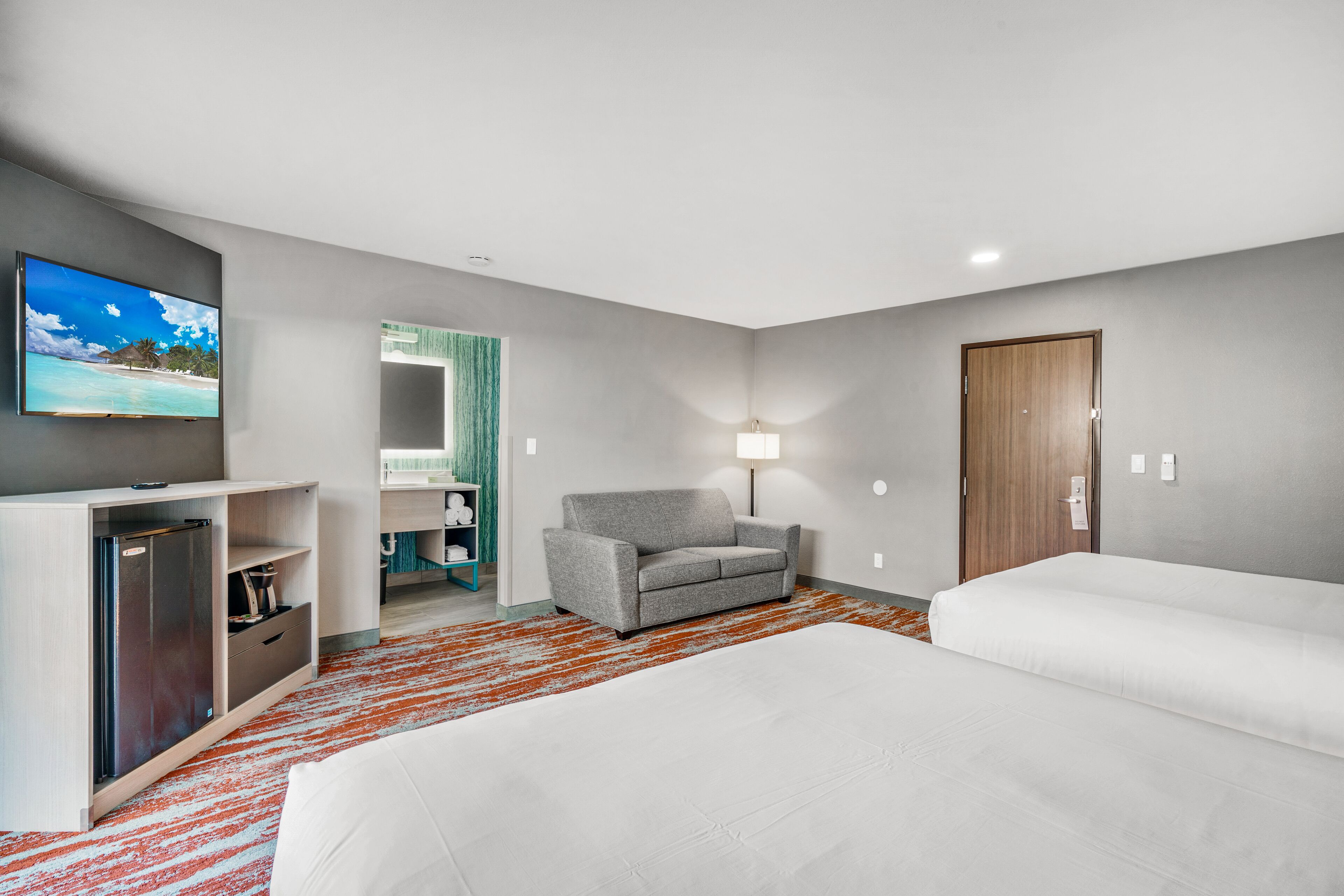 Hotelværelse med fjernsyn, sofa, to senge og et badeværelse, der ses gennem en åben dør.