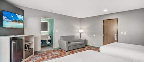 Hotelværelse med fjernsyn, sofa, to senge og et badeværelse, der ses gennem en åben dør.