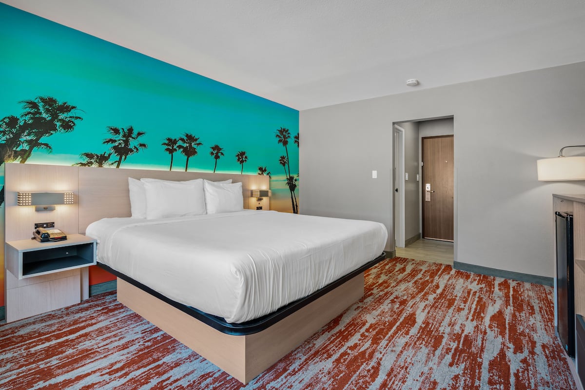 A modern hotel room with a large bed, a nightstand, a wall-mounted lamp, and a door leading to another room.