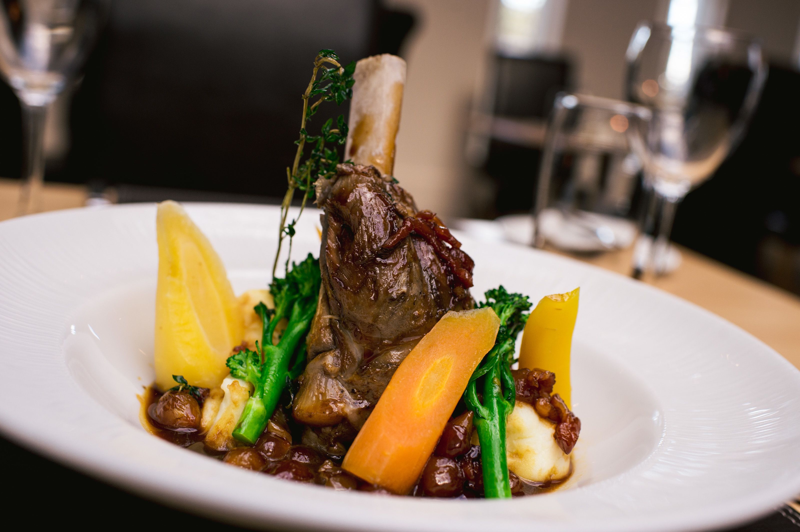 A plate of food with a lamb shank, vegetables, and a sauce.