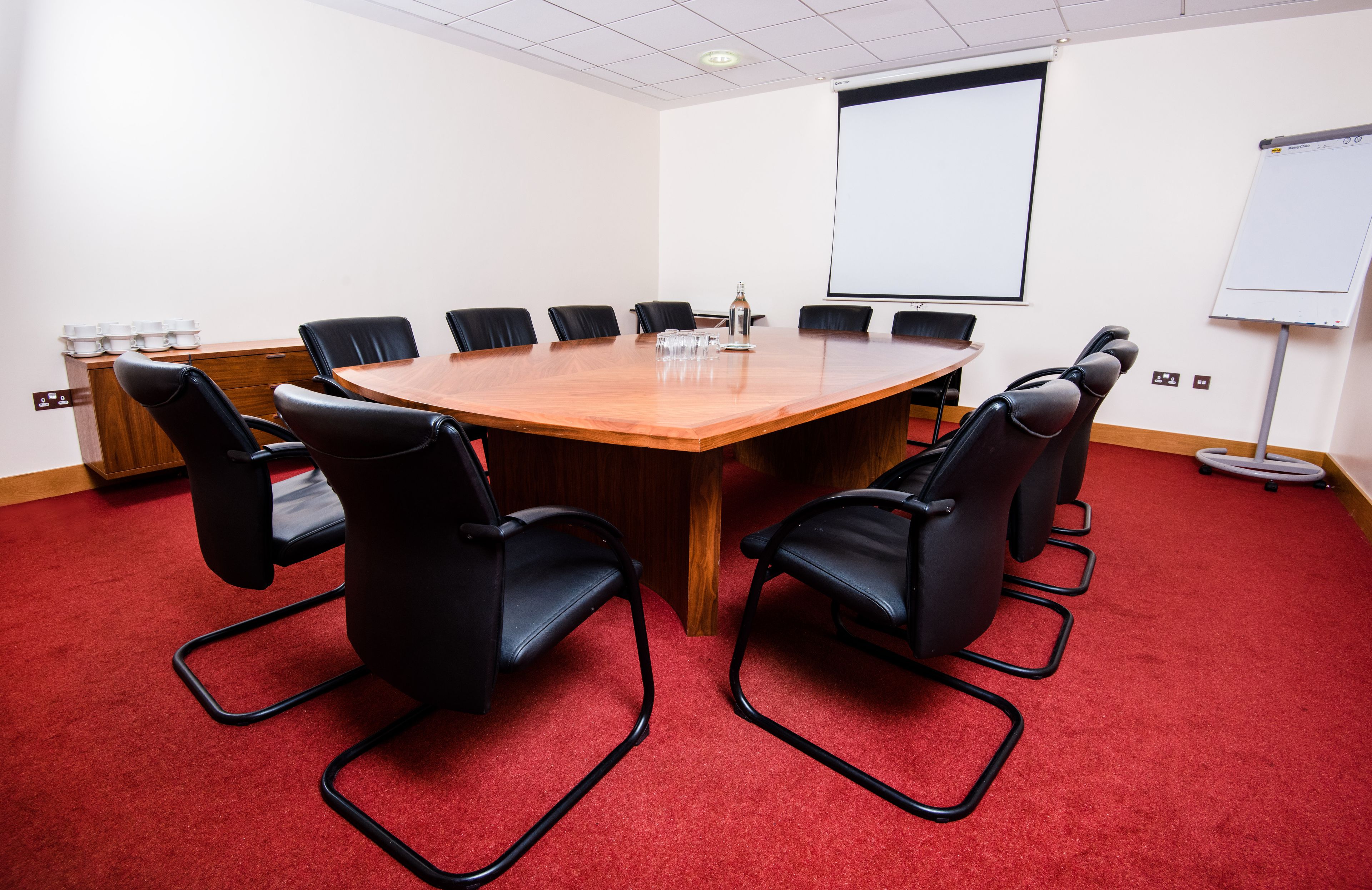 Una sala de conferencias con una mesa de madera, sillas negras, una pantalla de proyector y una pizarra.