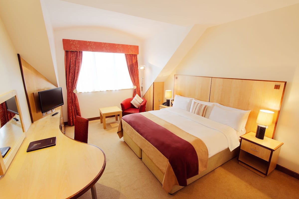 A hotel room with a bed, bedside table, lamp, TV, desk, chair, and a window with curtains.