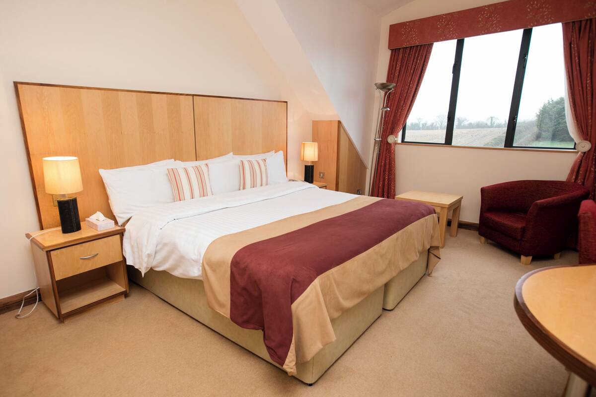 A hotel room with a large bed, bedside tables, a chair, and a window with curtains.