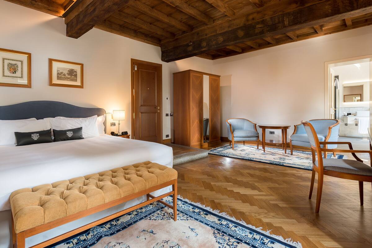 Ein Schlafzimmer mit einem großen Bett, einem Holz-Kopfteil und einer Bank. Im Raum befinden sich zwei Stühle und ein kleiner Tisch.