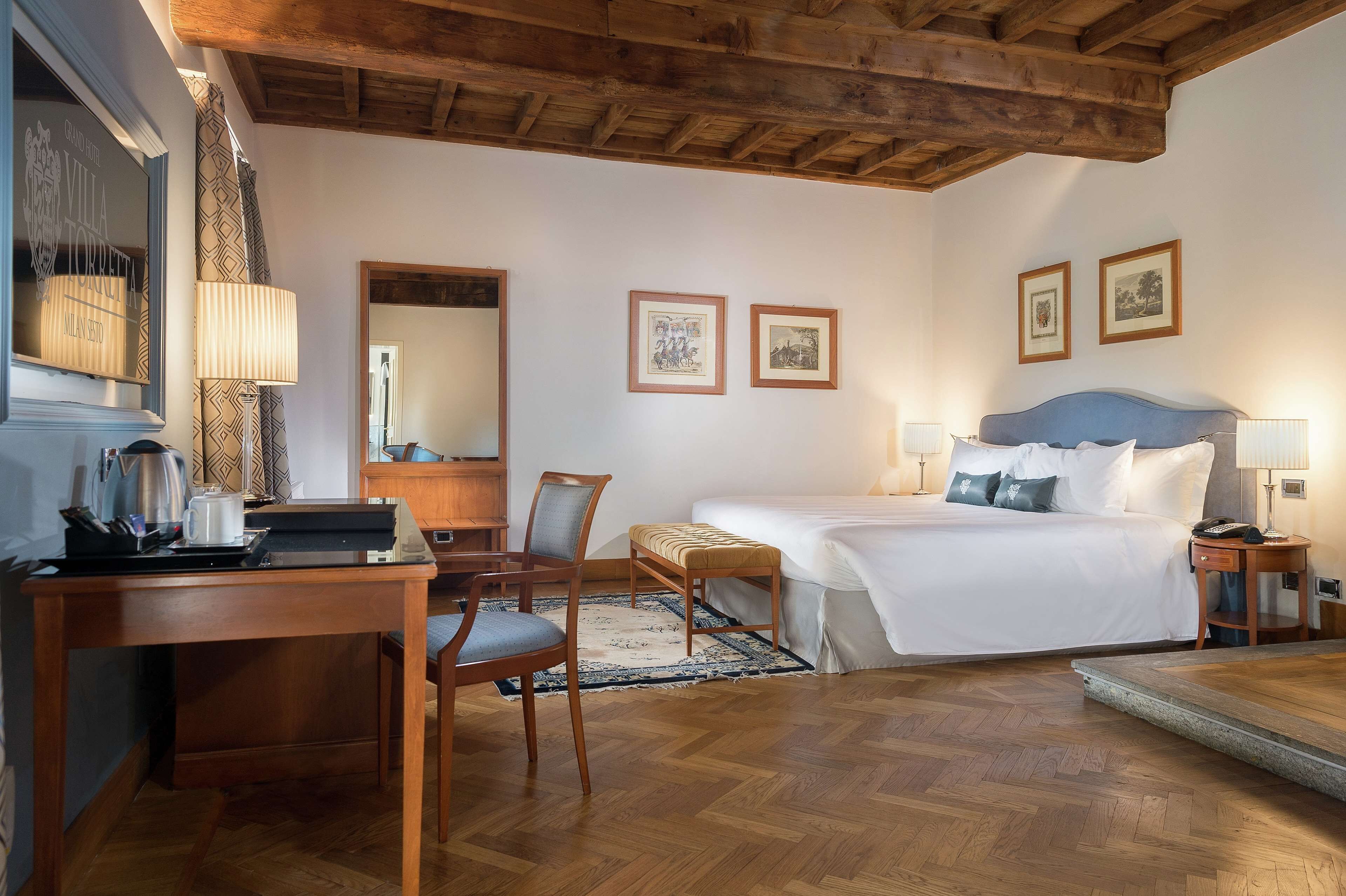 A hotel room with a wooden ceiling, a bed with white linens, a desk with a lamp, and two framed pictures on the wall.