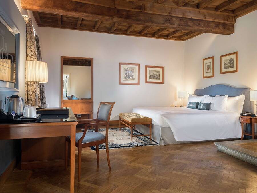 A hotel room with a wooden ceiling, a bed with white linens, a desk with a lamp, and two framed pictures on the wall.