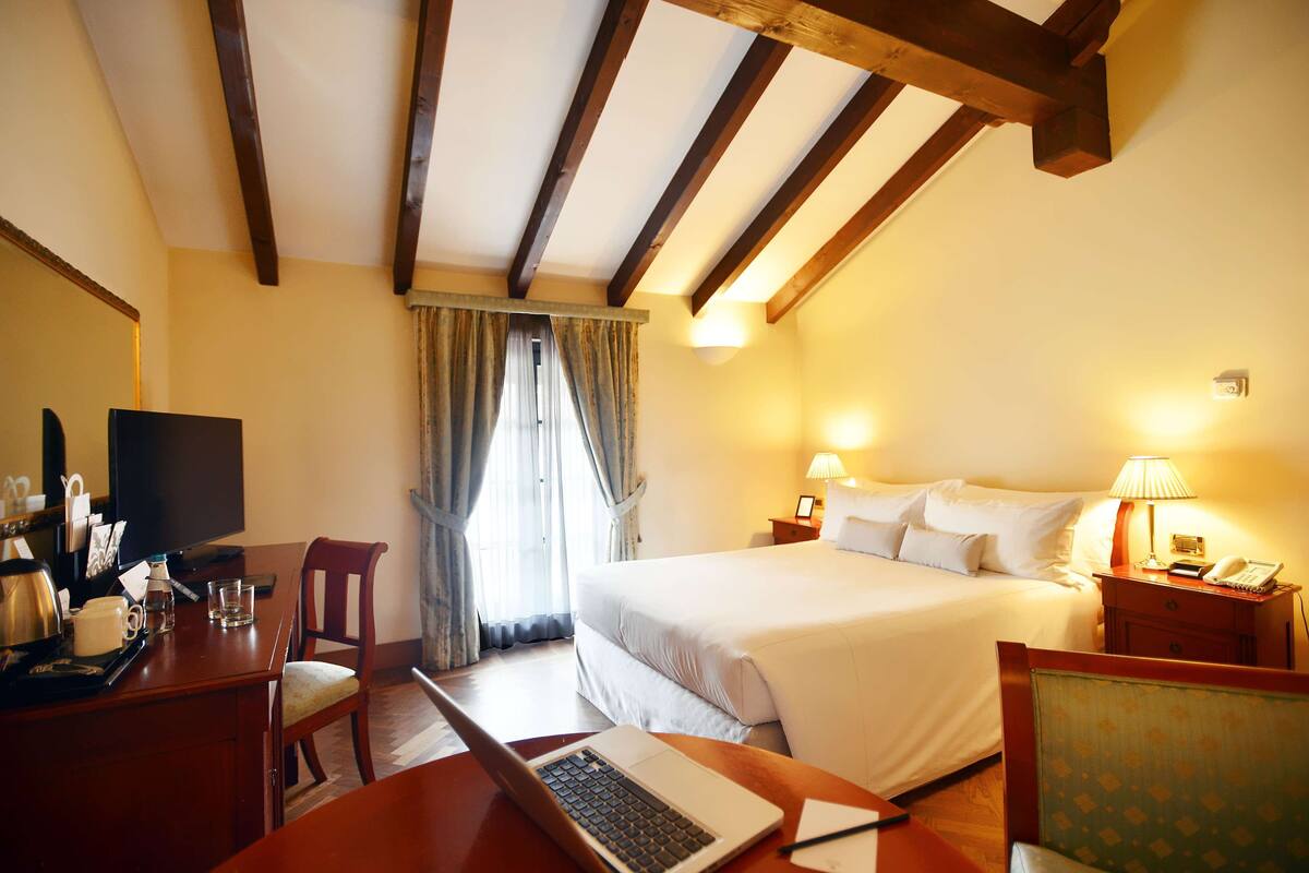 Ein Hotelzimmer mit einem Bett, einem Schreibtisch mit Laptop, einem Stuhl, einem Fernseher und einem Fenster mit Vorhängen.