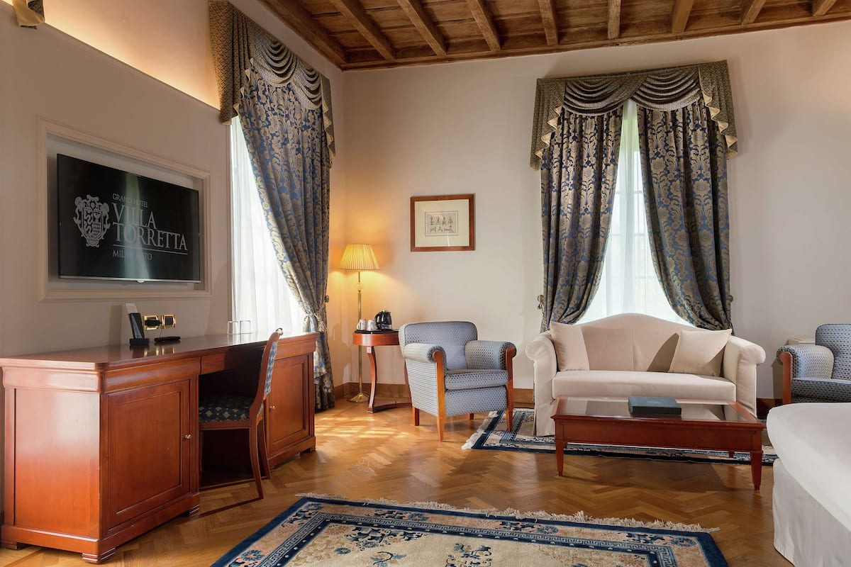 Ein Hotelzimmer mit einem Holztisch, einer Couch, einem Sessel und einem Bett. An der Wand hängt ein gerahmtes Bild und auf einem Nachttisch steht eine Lampe.