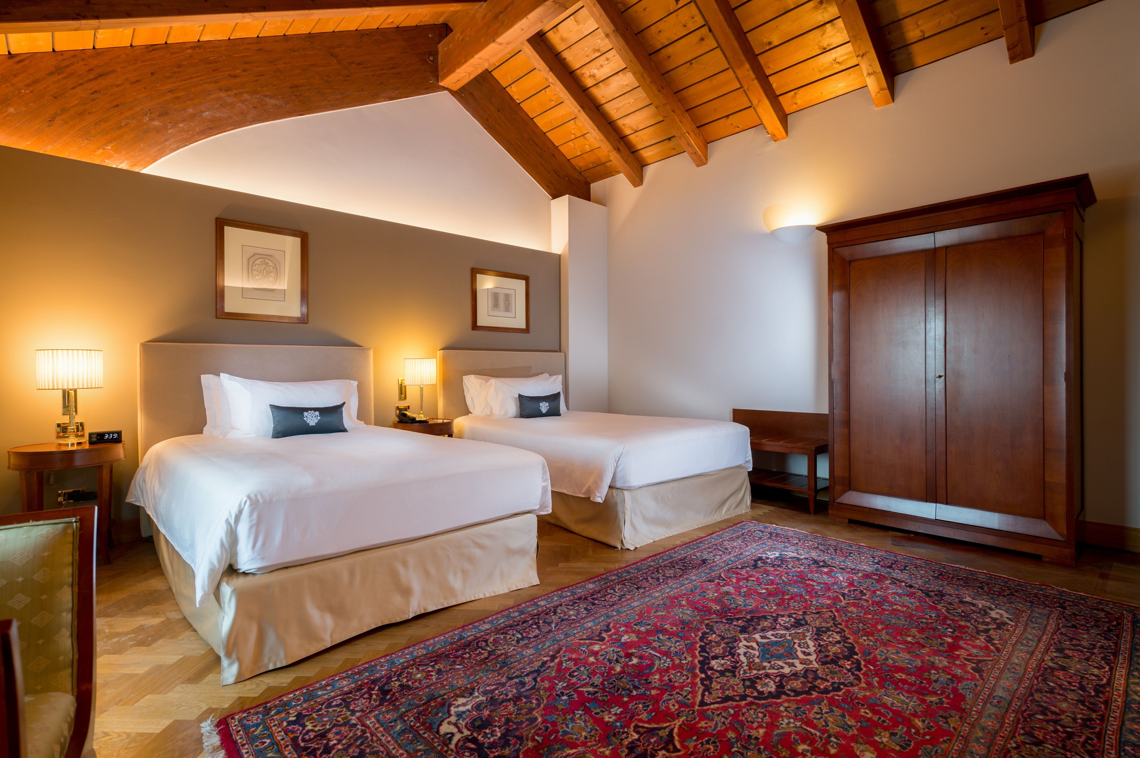 A hotel room with two beds, a large wooden wardrobe, a red patterned rug, and framed pictures on the wall.