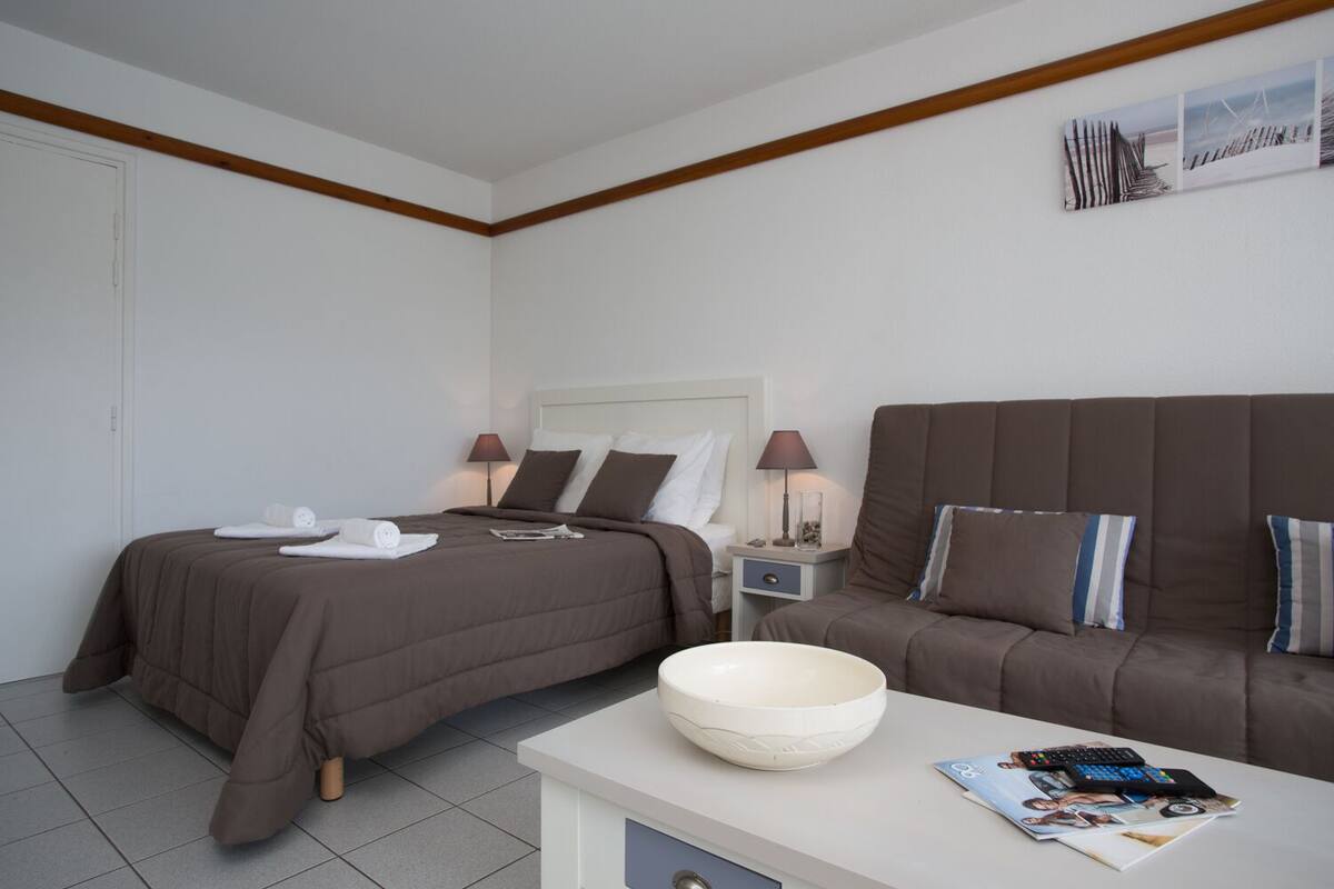 Ein Hotelzimmer mit einem Bett, einem Sofa, einem Nachttisch mit Lampen, einem weißen Tisch mit einer Schale und Zeitschriften sowie einer Wand mit gerahmten Bildern.