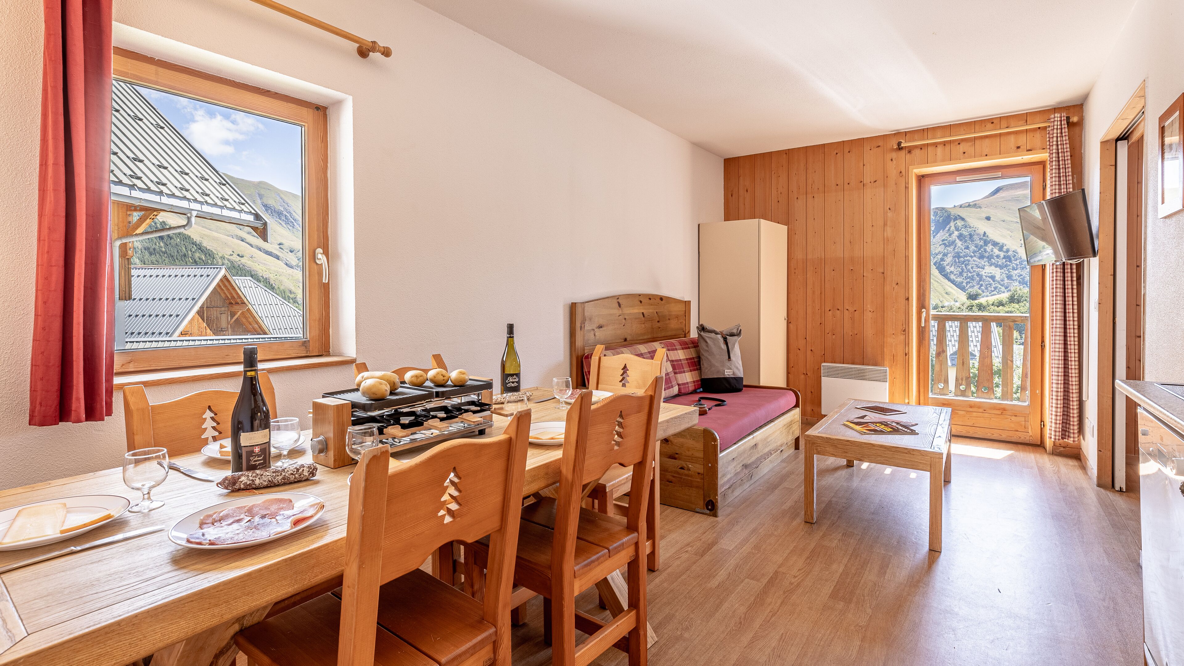 Une pièce chaleureuse avec une table à manger en bois, des chaises, un lit avec une tête de lit en bois, une petite table avec un livre, et une fenêtre donnant sur une montagne.