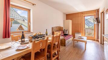 Une pièce chaleureuse avec une table à manger en bois, des chaises, un lit avec une tête de lit en bois, une petite table avec un livre, et une fenêtre donnant sur une montagne.
