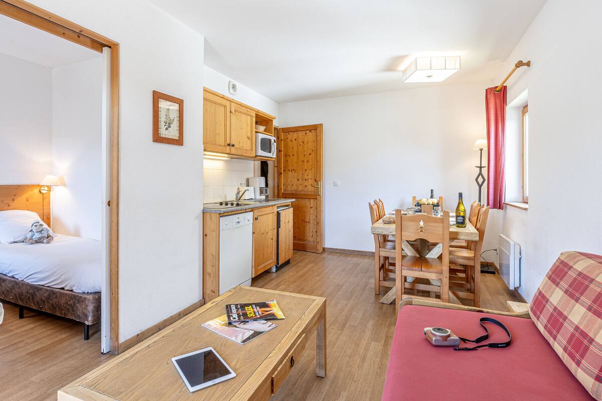 Et kompakt værelse med seng, et lille bord med en tablet, et træspisebord med stole, et tekøkken med træskabe og en rød sofa.