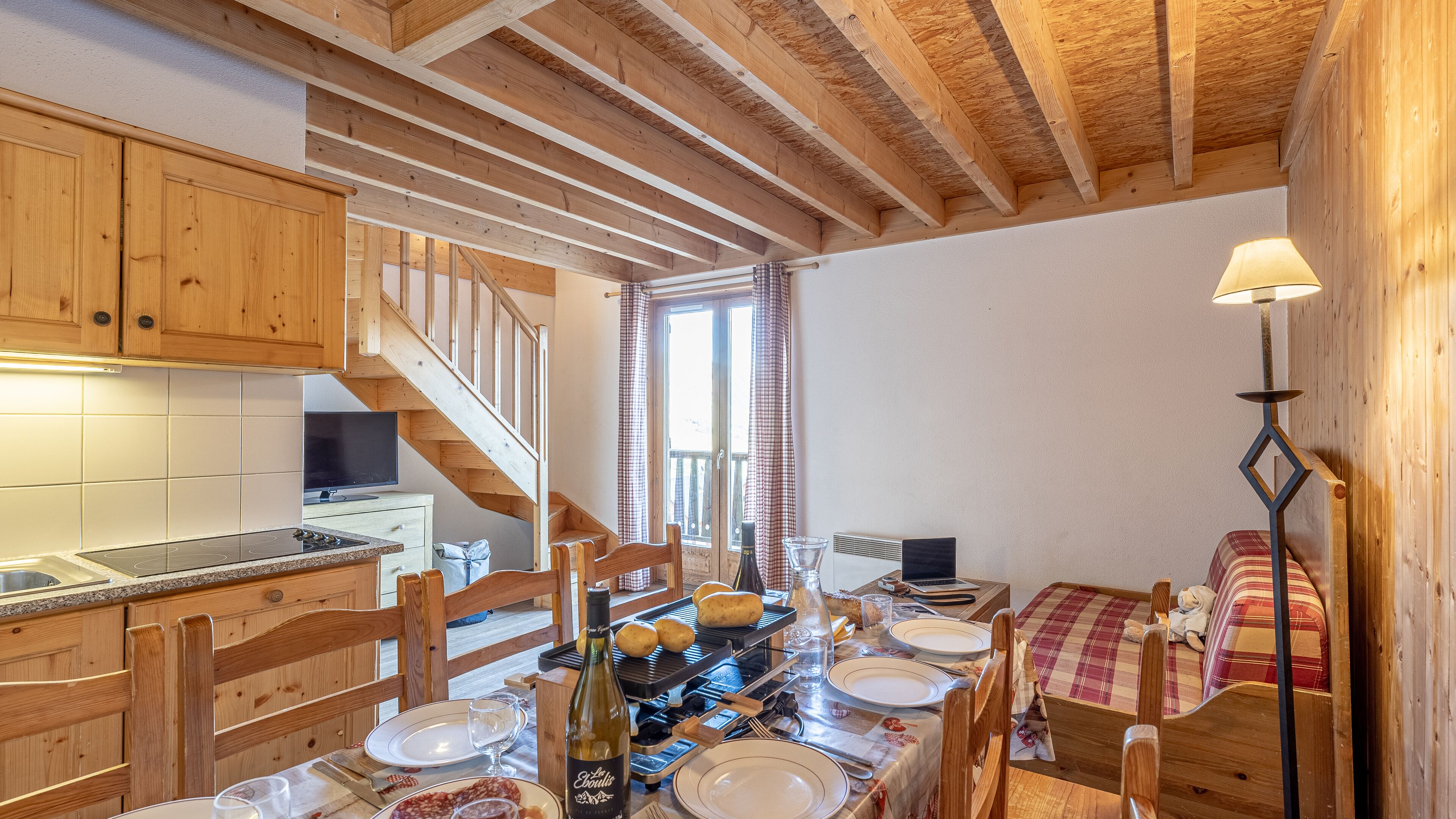 Une cuisine chaleureuse avec des meubles en bois, une table à manger dressée pour deux personnes et un escalier menant à un balcon.