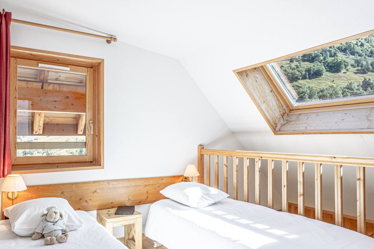 Et soveværelse med sengegavl i træ, et vindue med rødt gardin, en seng med hvide sengetæpper, et natbord med lampe, og et billede i ramme på væggen.