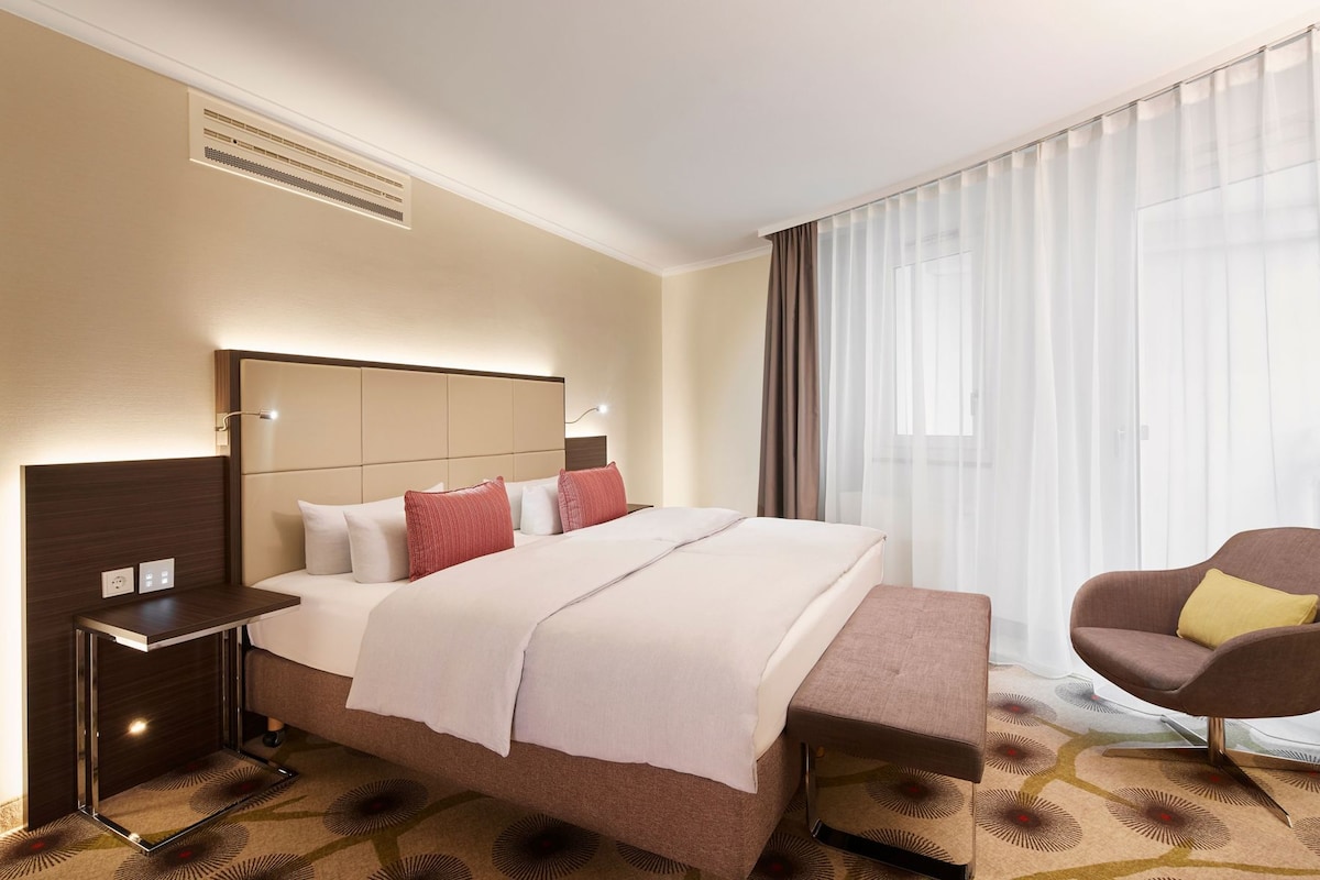 Habitación de hotel con una cama grande, mesitas de noche, una silla y un ventanal con cortinas translúcidas.