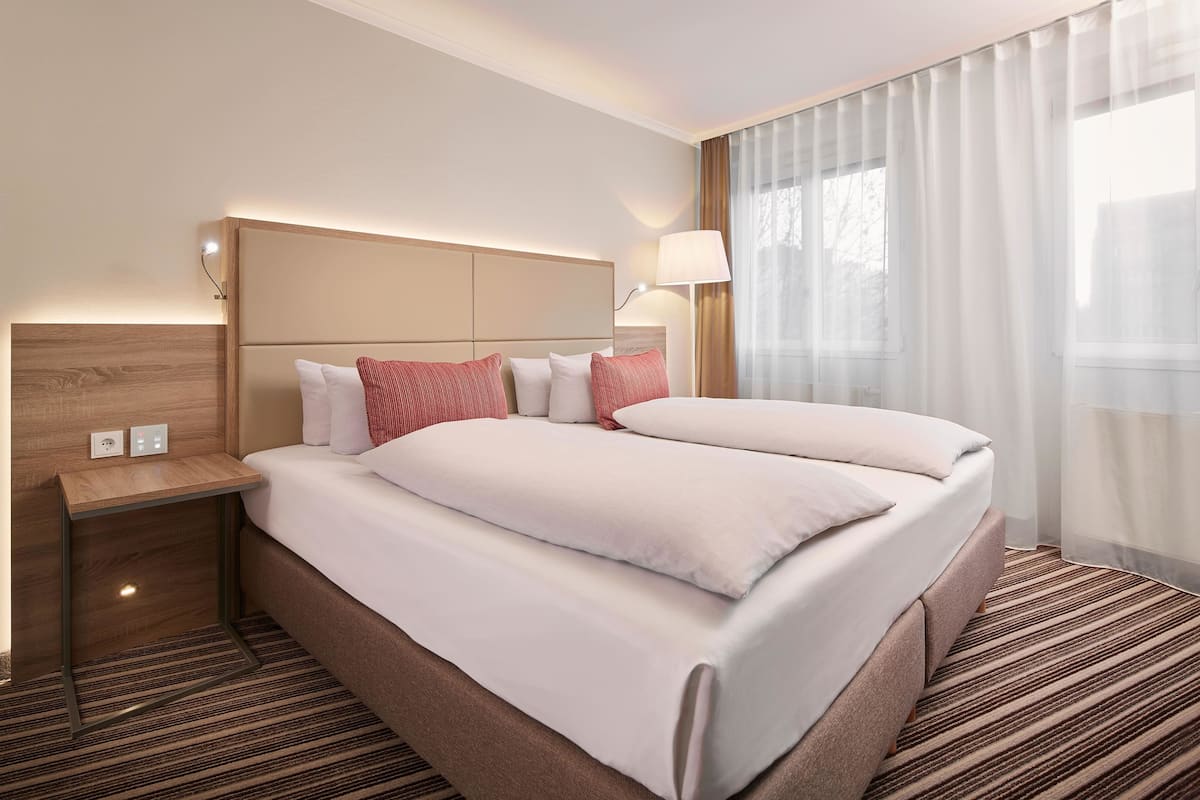 A hotel room with a large bed, a bedside table, a lamp, and a view of the outdoors through sheer curtains.