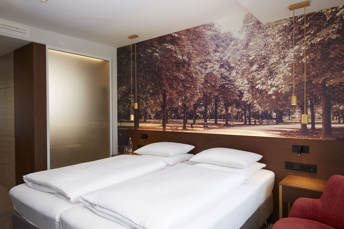 A hotel room with a large bed, a red chair, a nightstand, and a wall mural of trees.