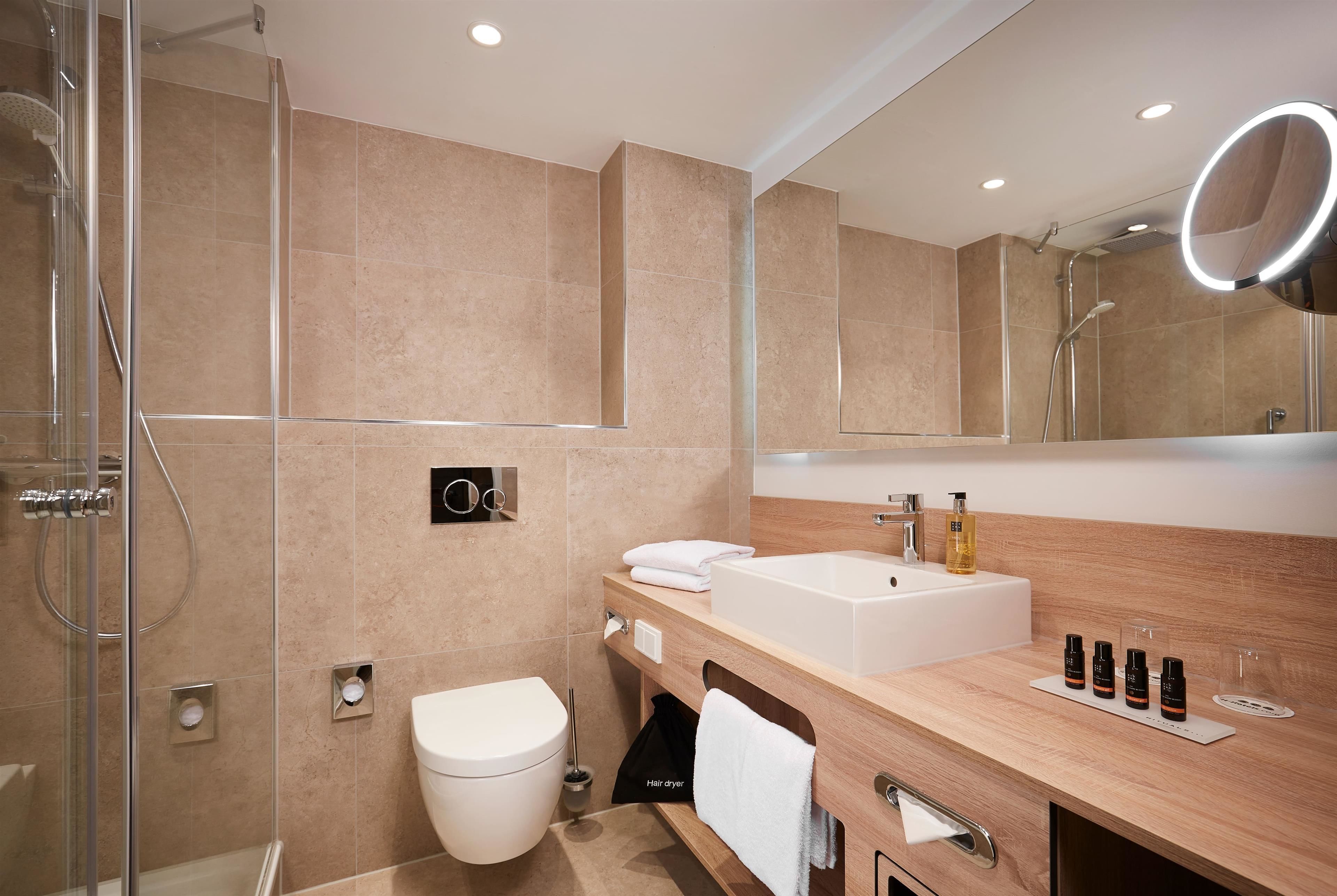 Un baño moderno con cabina de ducha de vidrio, inodoro blanco, tocador de madera con lavamanos y espejo con luz redonda.