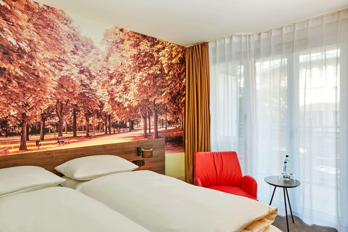 A hotel room with a bed, a red chair, a small table with a bottle and glasses, and a mural of a park with trees.