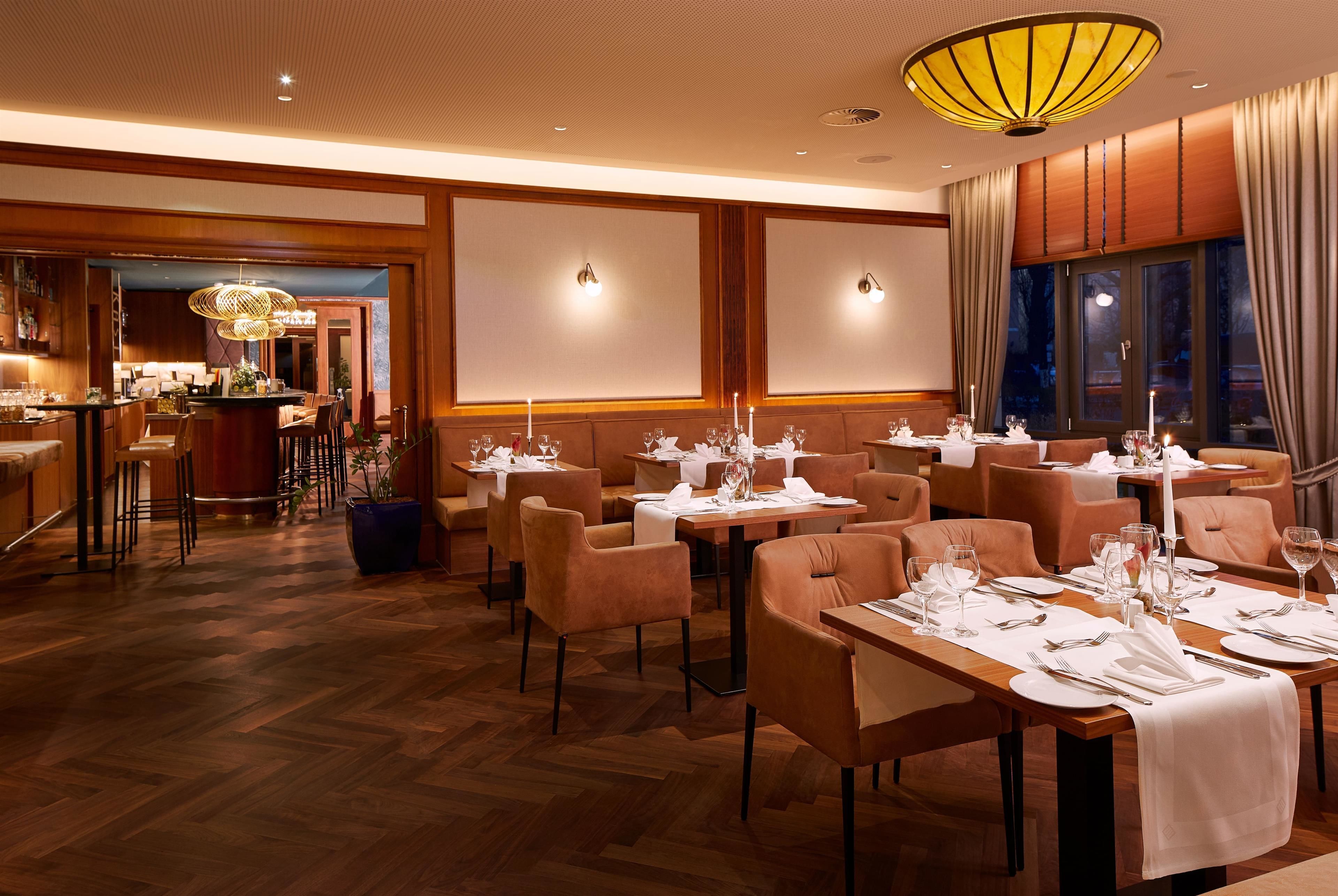 Un restaurante con piso de madera, varias mesas de comedor dispuestas con manteles blancos y velas, y una zona de bar visible al fondo.
