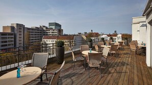 A rooftop terrace with wicker chairs and tables, potted plants, and a view of city buildings. - HYPERION Hotel Berlin (Berlin)