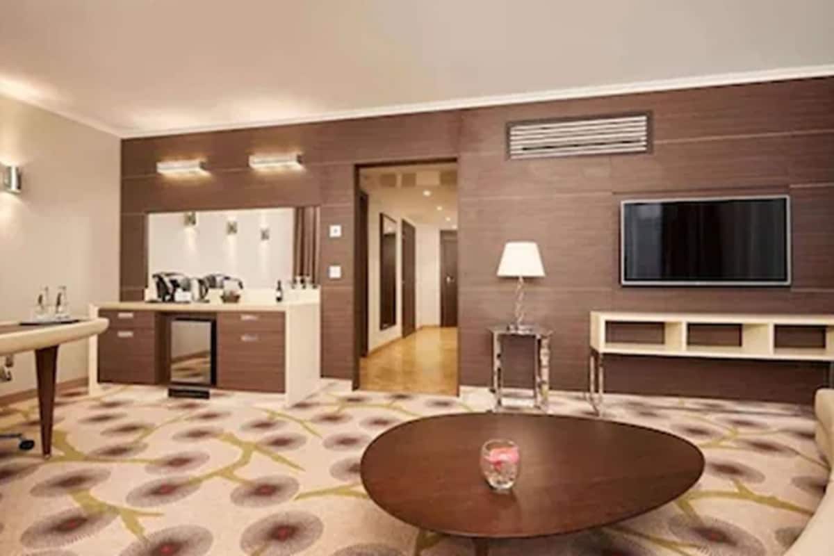 A modern hotel room with a large bed, a round coffee table, a TV mounted on the wall, and a small side table with a lamp.