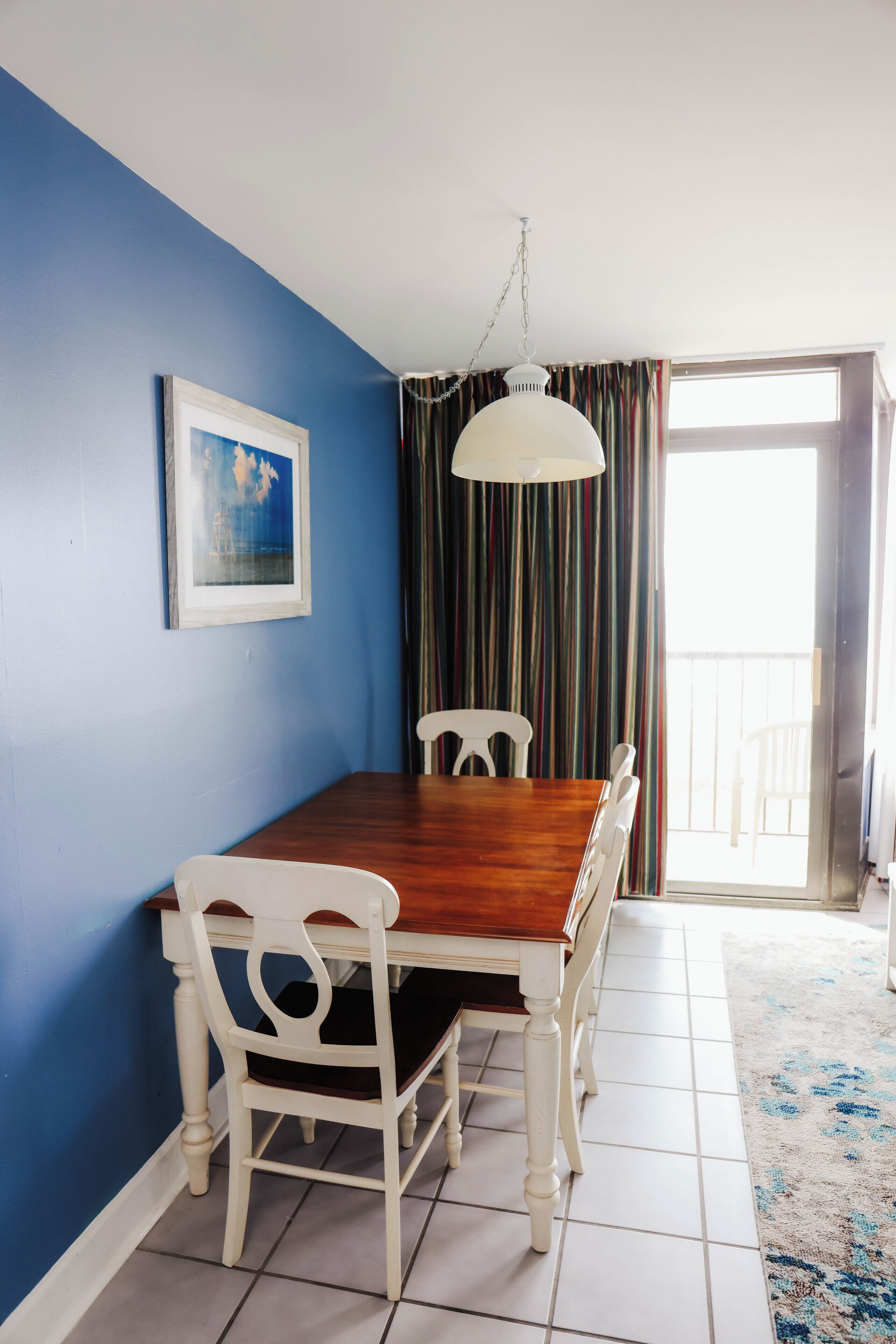 Un coin repas avec une table en bois et des chaises blanches, un tableau encadré au mur et une vue sur l’extérieur par une porte.