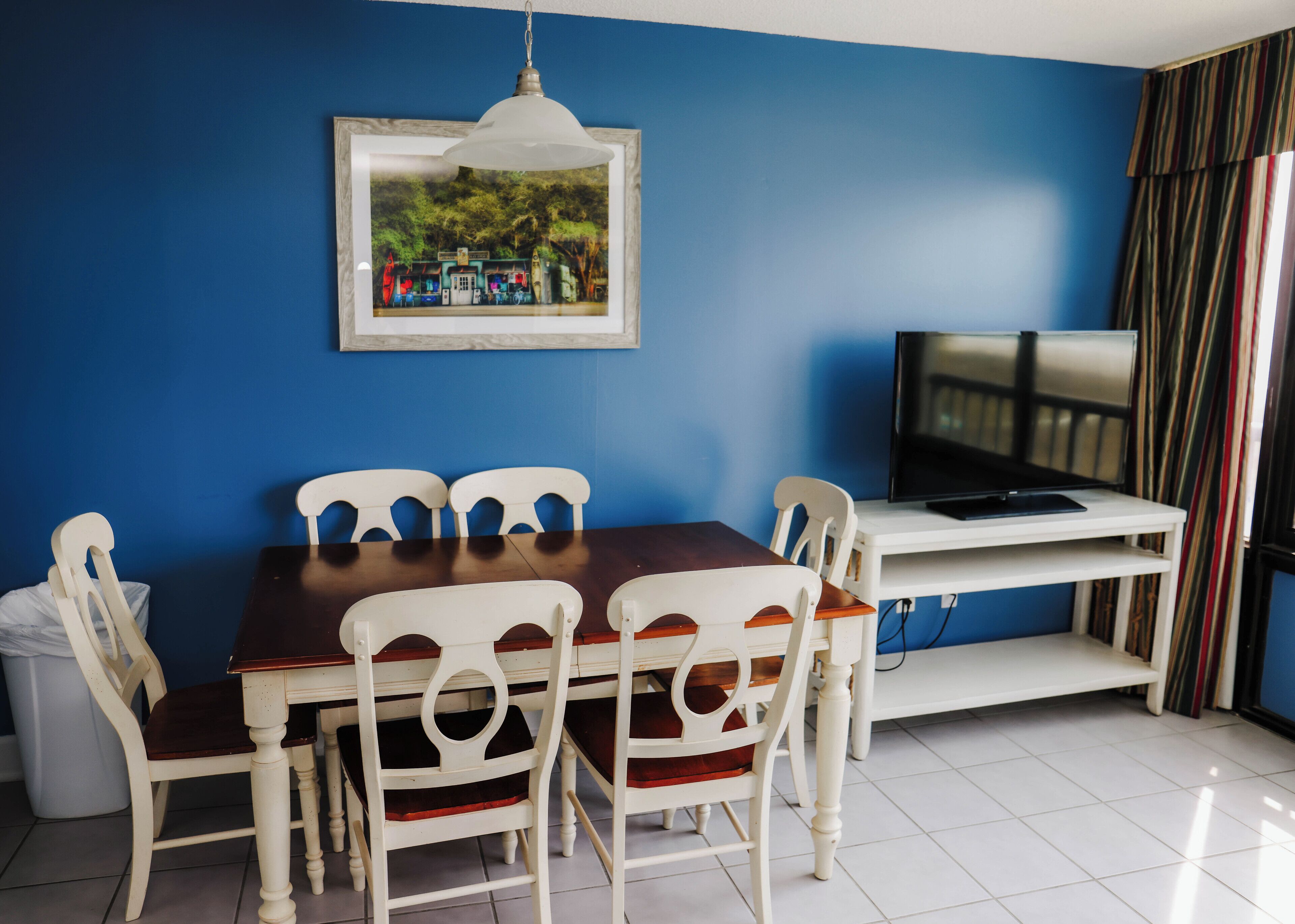 A dining area with a wooden table and white chairs, a television on a stand, and a framed picture on the wall.