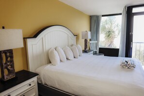 A hotel room with a white bed, a nightstand with a lamp, and a view of palm trees through the window.