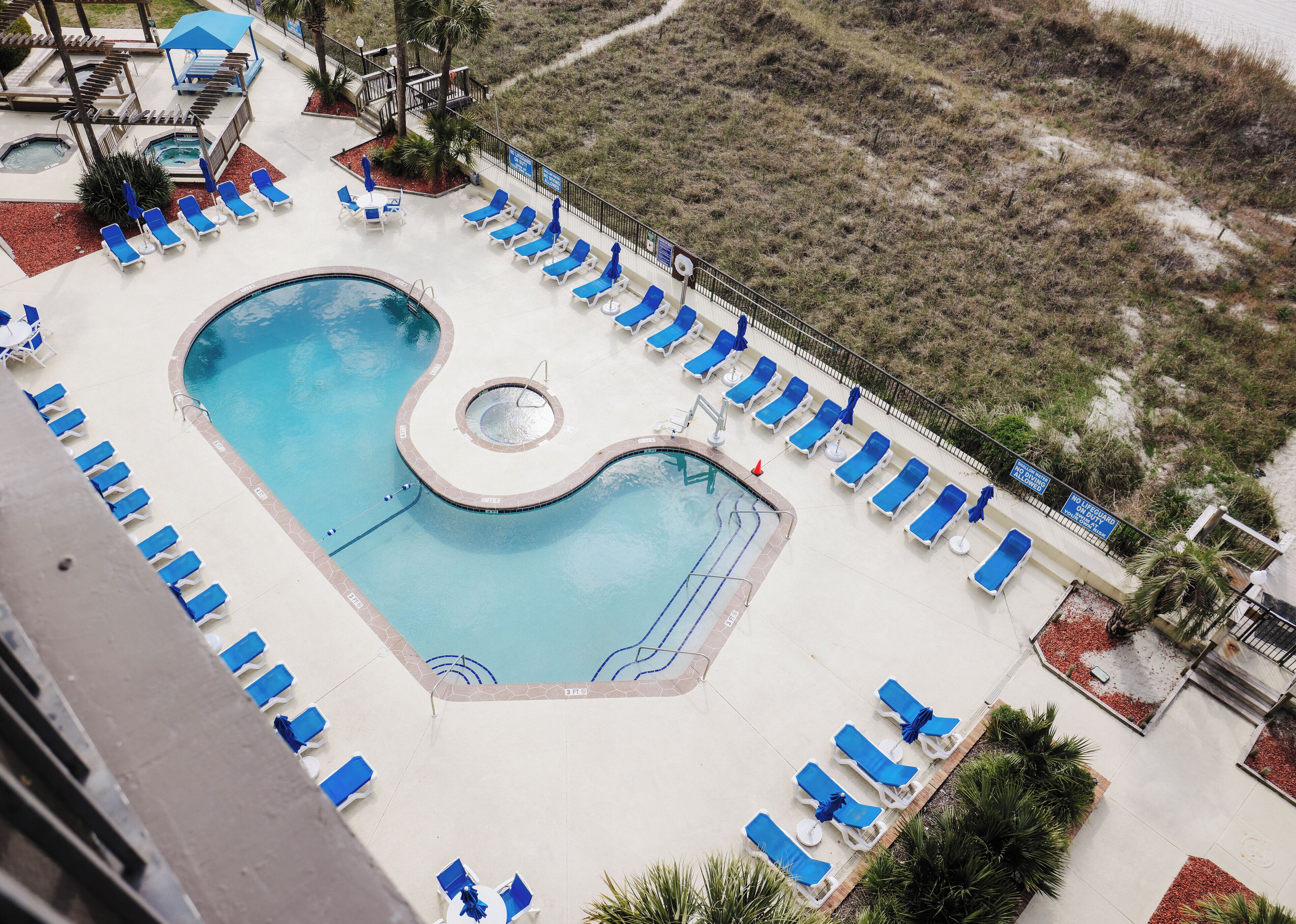 A swimming pool with a unique design, surrounded by blue lounge chairs and umbrellas, with a view of a grassy area in the background.
