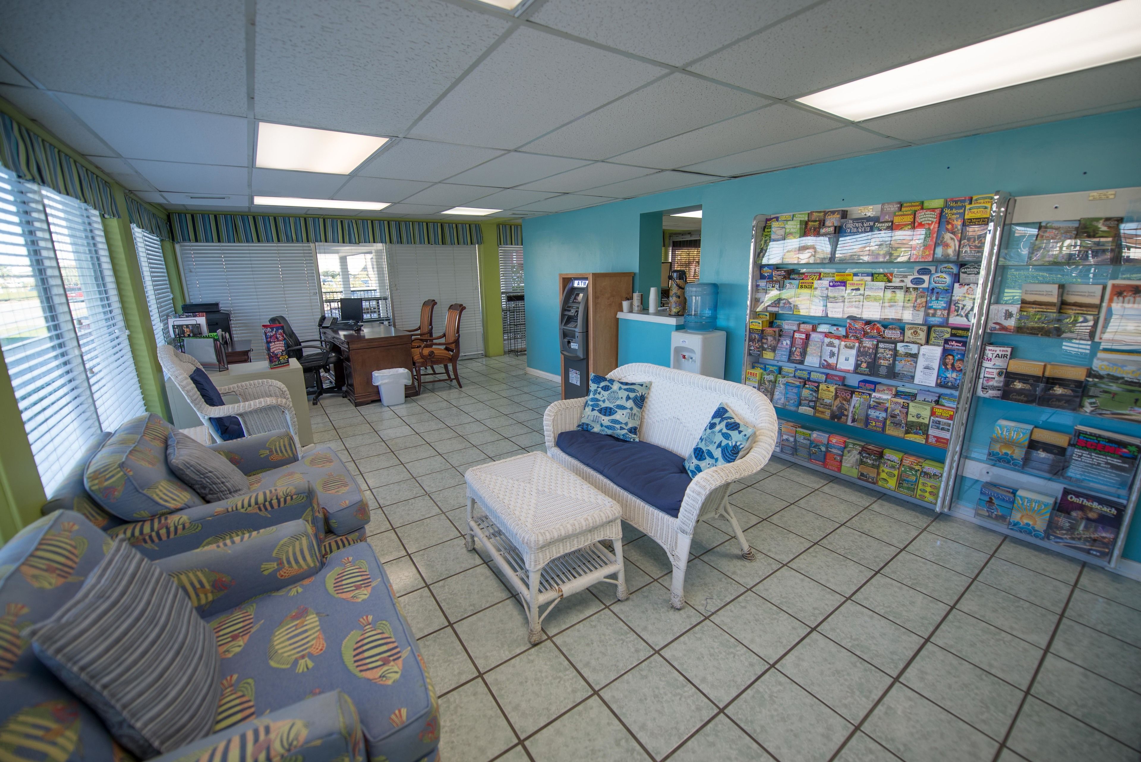 A room with a reception desk, a computer, a water dispenser, a magazine rack, and a seating area with patterned sofas and a small table.