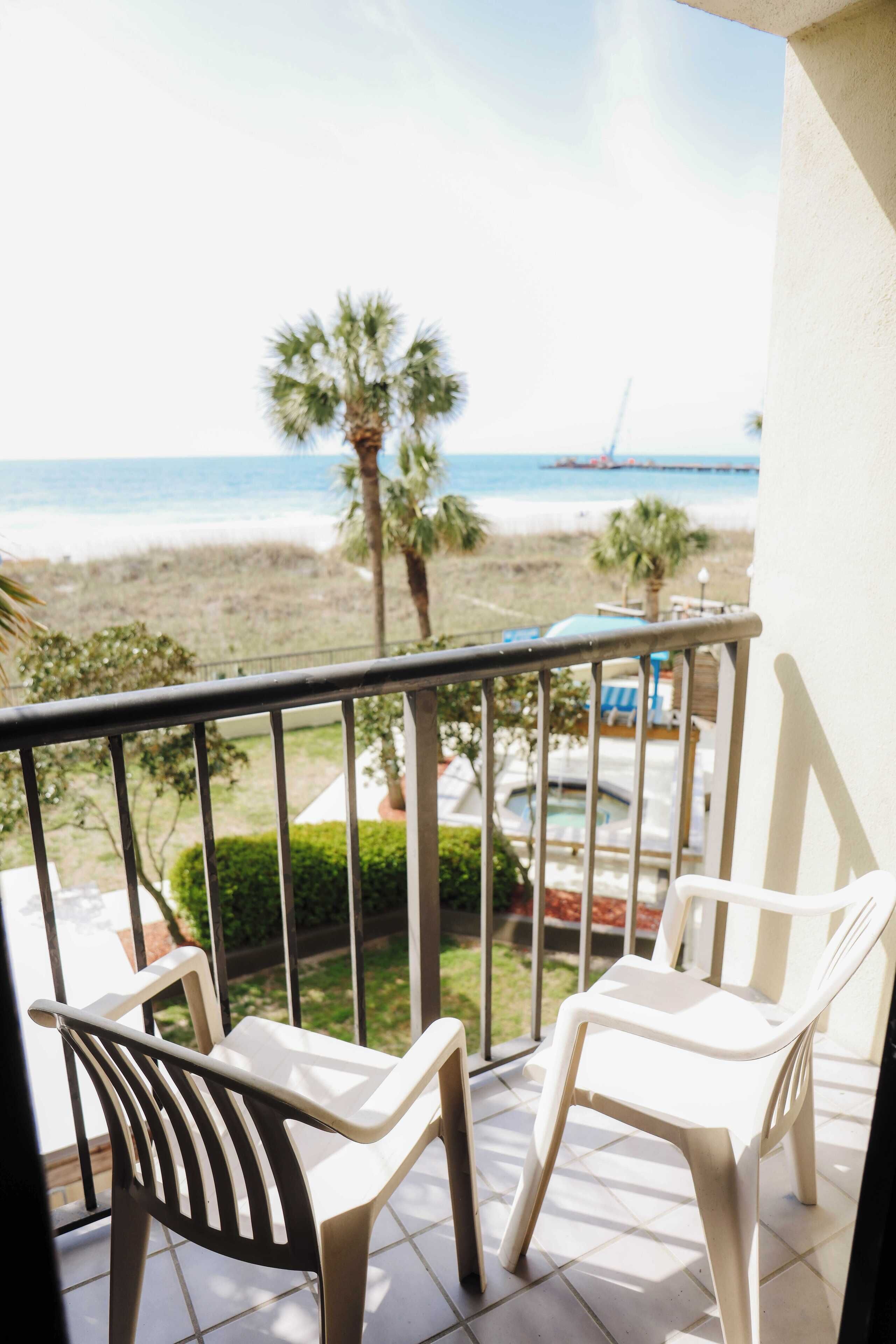 A balcony with two white plastic chairs, a view of the beach, and a pool area in the background.